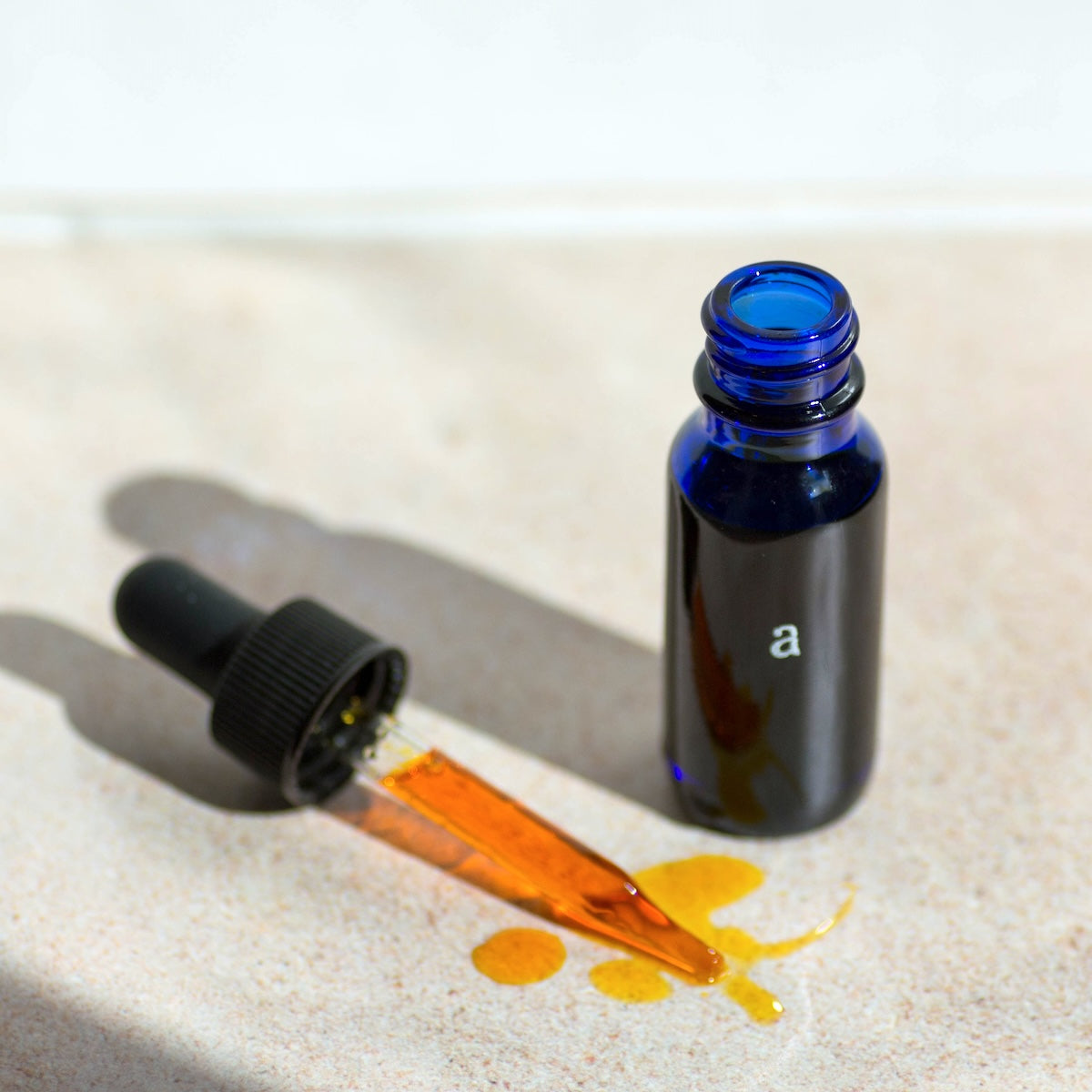 A half-ounce Age Care Facial Serum in a vibrant blue glass dropper bottle with visible dropper spilling a golden, fast-absorbing oil onto a sunlit, sandy surface, embodying the nourishing blend of Sea Buckthorn Oil, Squalane from Olives, and Frankincense Essential Oil with CoQ10 for anti-aging and moisturizing, ideal for all skin types, vegan from our facial care collection.
