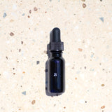 Age Care Facial Serum in a .5 oz elegant blue glass bottle with a black dropper, prominently featuring the letter 'a' in white text, poised on a coarse sandy backdrop sprinkled with small pebbles, highlighting the product's vegan, fast-absorbing oil formula suitable for all skin types and designed to combat aging and dryness, from our comprehensive facial care collection.