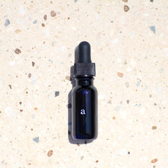 Age Care Facial Serum in a .5 oz elegant blue glass bottle with a black dropper, prominently featuring the letter 'a' in white text, poised on a coarse sandy backdrop sprinkled with small pebbles, highlighting the product's vegan, fast-absorbing oil formula suitable for all skin types and designed to combat aging and dryness, from our comprehensive facial care collection.